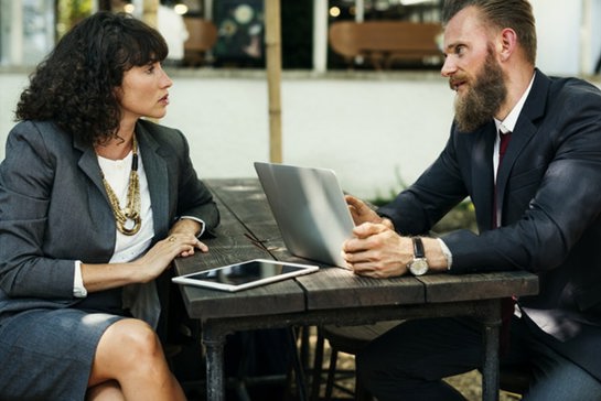 Two business professionals having a serious discussion outdoors with a laptop and tablet on a wooden table.