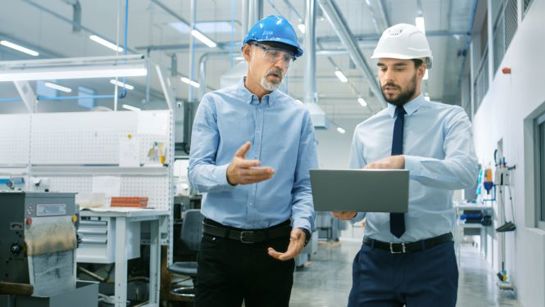 Two engineers wearing hard hats discussing work in a modern factory while reviewing data on a laptop.