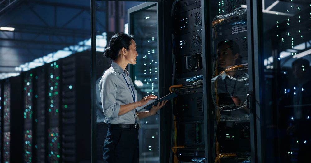 Female Network Technician Standing With Digital Tablet Computer In Front Of Open Server Rack Cabinet In Data Centre