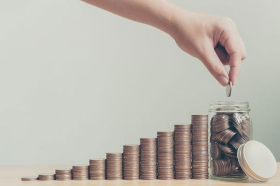 Hand placing a coin in a jar beside stacked coins arranged in ascending order, symbolising savings or growth.