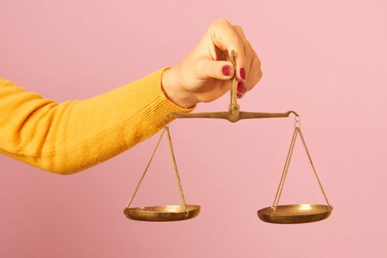 Hand holding a brass balance scale against a pink background, symbolizing justice, fairness, or decision-making.