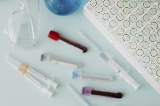 Lab equipment with test tubes, syringes, a microplate, and safety glasses on a light blue surface, for medical research