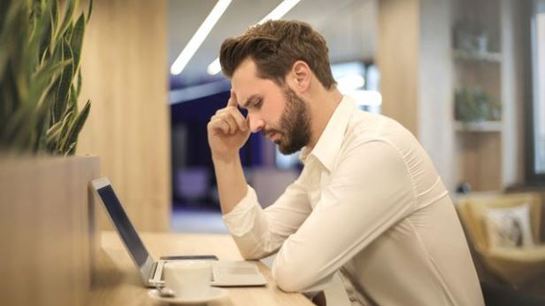 Stressed man in white shirt working on a laptop at a cafe, resting his head on his hand in frustration.